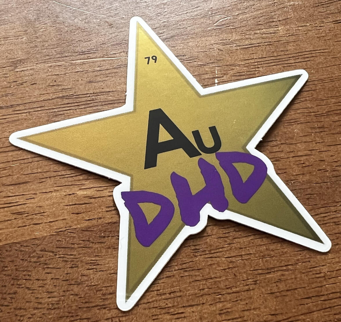 Photograph of a metallic gold sticker in the shape of a five-pointed star. The top point of the star has the Periodic Table number 79 on it, in the same font as used in the table, and some text below in the same font reads "Au," the abbreviation for gold i