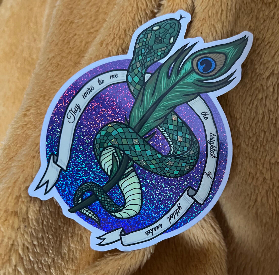 A photograph of a sticker featuring a circular sparkly purple background, upon with sits an image of a green snake coiling around a peacock feather. The snake has some gold sparkly scales scattered along its body, and the image is encircled by a Medieval s